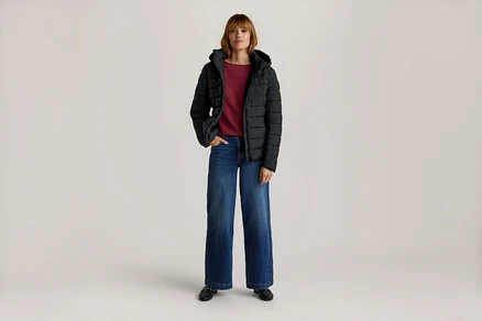 Frau steht in schwarzer Kapuzen-Steppjacke, rotem Langarmshirt, blauer weiter Jeans und schwarzen Slippern, eine Hand in der Tasche.
