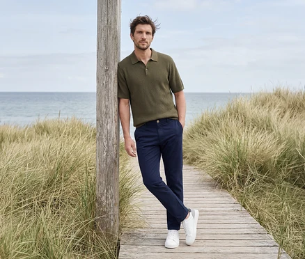 Mann in olivgrünem Poloshirt, dunkelblauer Hose und weißen Sneakers lehnt an einem Holzpfosten am Strandweg mit Meer im Hintergrund.