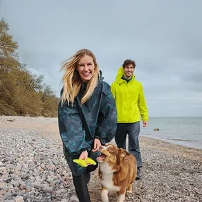 Eine lächelnde Frau in einem dunklen Regenponcho hält einen gelben Frisbee, während sie mit einem braun-weißen Hund an einem Kiesstrand spaziert. Ein lächelnder Mann in einer gelben Regenjacke geht hinter ihnen, mit Meer und bewölktem Himmel im Hintergrund.