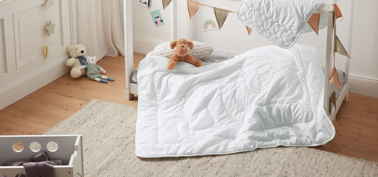 Ein Kinderbett mit weißem Rahmen, dekoriert mit Wimpeln, mit einer weißen irisette® greenline Kinder-Steppdecke und einem Kissen. Auf dem Bett liegt ein Teddybär.
