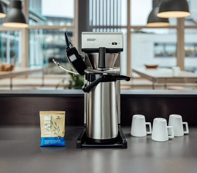 Auf einem Tisch stehen eine Profi-Filterkaffeemaschine »Bravilor-Bonamat TH« ohne Kanne, drei Tassen und eine Packung Kaffee.