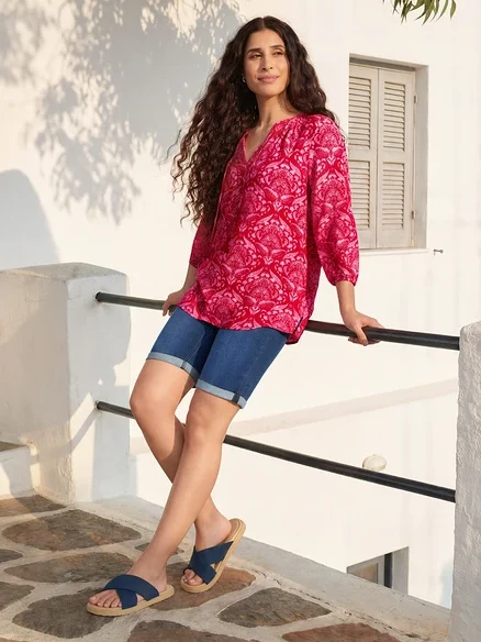 Frau lehnt an Geländer, trägt pinke Tunika-Bluse, Bermuda-Jeans und Komfortsandalen. Sie trägt Creolen Pavé.