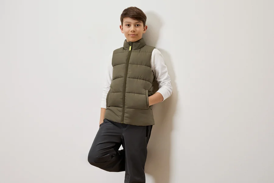 Ein Junge mit braunen Haaren lehnt an einer weißen Wand und trägt eine olivgrüne Steppweste über einem weißen Langarmshirt und graue Hosen.