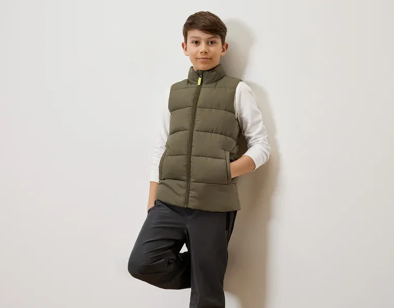 Ein Junge mit braunen Haaren lehnt an einer weißen Wand und trägt eine olivgrüne Steppweste über einem weißen Langarmshirt und graue Hosen.