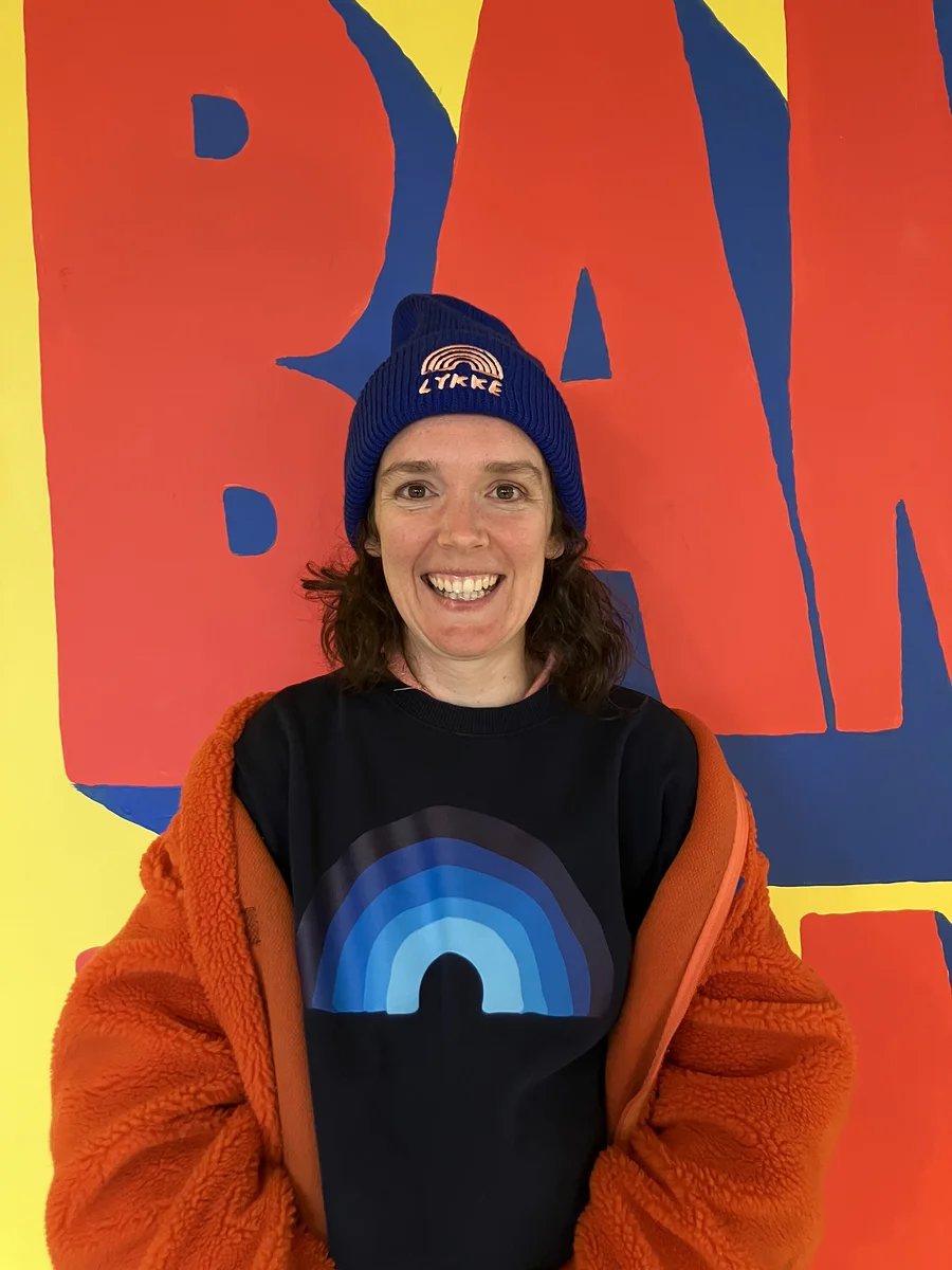Eine lächelnde Frau mit dunklen Haaren blickt in die Kamera. Sie trägt eine blaue Mütze mit „LYKKE“-Schriftzug und Regenbogen, ein dunkelblaues Sweatshirt mit blauem Regenbogenmotiv und eine orangefarbene flauschige Jacke über den Schultern. Der Hintergrund ist ein farbenfrohes Wandgemälde mit großen roten Formen auf gelbem und blauem Grund.