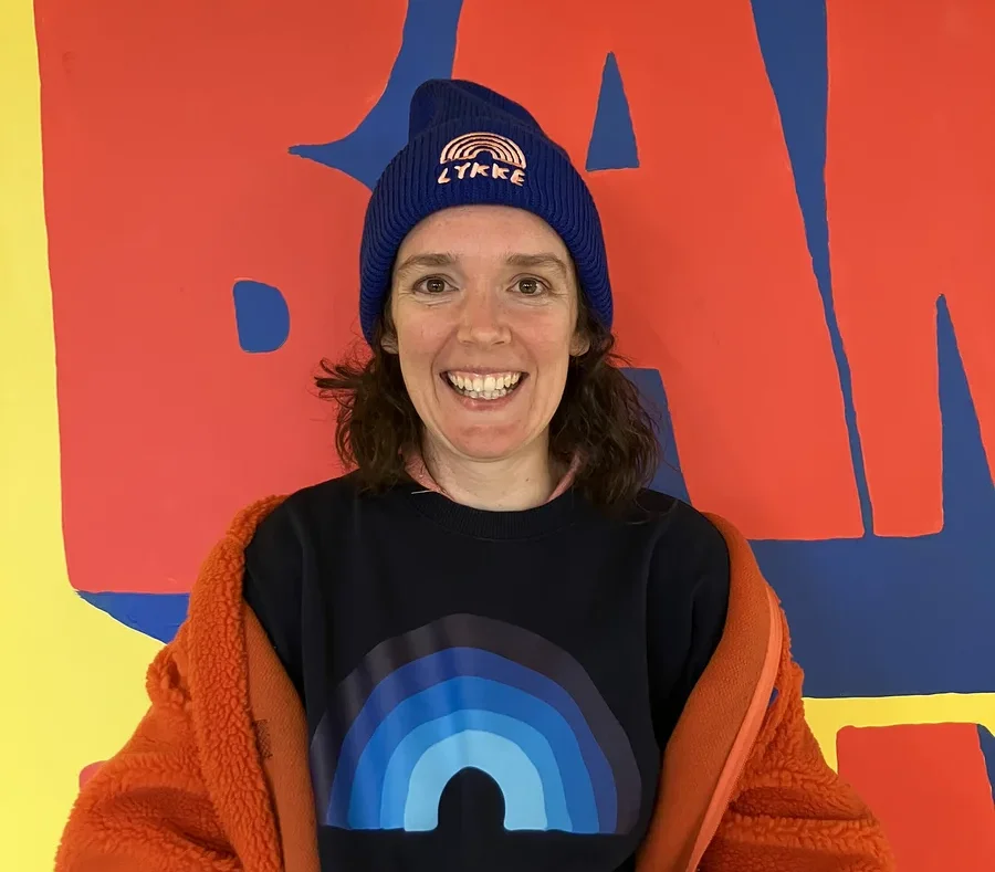 Eine lächelnde Frau mit dunklen Haaren blickt in die Kamera. Sie trägt eine blaue Mütze mit „LYKKE“-Schriftzug und Regenbogen, ein dunkelblaues Sweatshirt mit blauem Regenbogenmotiv und eine orangefarbene flauschige Jacke über den Schultern. Der Hintergrund ist ein farbenfrohes Wandgemälde mit großen roten Formen auf gelbem und blauem Grund.