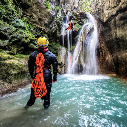 Eine Person in schwarzem Neoprenanzug, gelbem Helm und orangefarbenem Seil steht im türkisfarbenen Wasser und beobachtet eine andere Person, die sich einen Wasserfall abseilt. Zwei weitere sind auf moosbewachsenen Felsen oben.