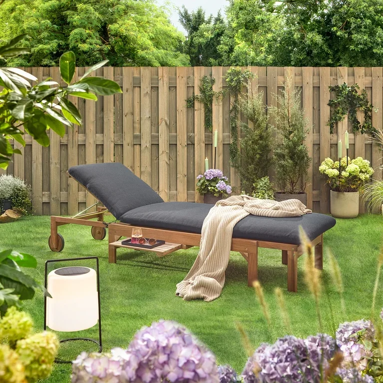 Gartenszene mit einer hölzernen Liege mit schwarzer Polsterung, einer Decke, einem Getränk, einer Sonnenbrille und einer Laterne auf einem Rasen vor einem Holzzaun.