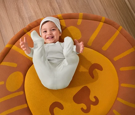 Ein lächelndes Baby liegt auf einem Kinderzimmer-Teppich »Löwe« und trägt ein 3-teiliges Baby-Set.