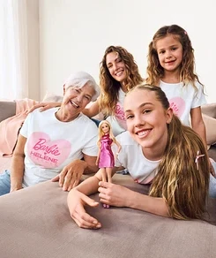 Vier lächelnde Frauen verschiedener Generationen, alle in weißen Barbie Helene Fischer T-Shirts, posieren auf einem grauen Sofa. Eine ältere Frau trägt hellblaue Palazzo Jeans – Fit »Neni«. Ein jüngeres Mädchen hält eine Barbie-Puppe.