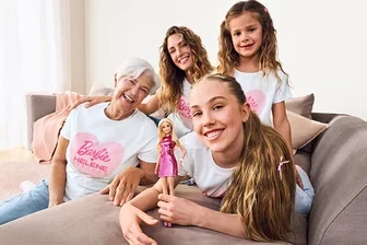 Vier lächelnde Frauen verschiedener Generationen, alle in weißen Barbie Helene Fischer T-Shirts, posieren auf einem grauen Sofa. Eine ältere Frau trägt hellblaue Palazzo Jeans – Fit »Neni«. Ein jüngeres Mädchen hält eine Barbie-Puppe.