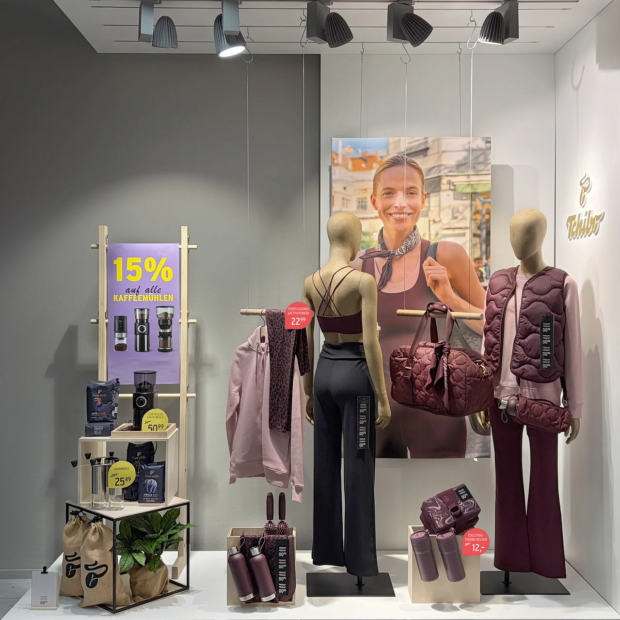 Schaufensterauslage mit zwei Schaufensterpuppen in bordeauxroter Sportkleidung, einem Poster einer lächelnden Frau, Kaffeemühlen und Kaffee. Dazu passende bordeauxrote Accessoires wie Reisetasche und Trinkflaschen. Ein Schild bewirbt 15% Rabatt auf Kaffeemühlen.