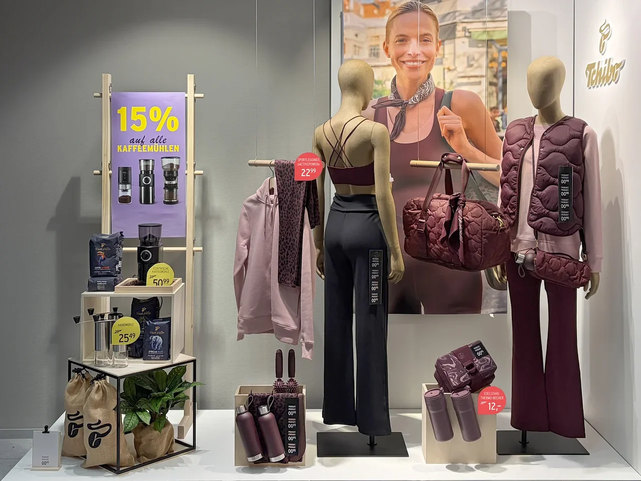 Schaufensterauslage mit zwei Schaufensterpuppen in bordeauxroter Sportkleidung, einem Poster einer lächelnden Frau, Kaffeemühlen und Kaffee. Dazu passende bordeauxrote Accessoires wie Reisetasche und Trinkflaschen. Ein Schild bewirbt 15% Rabatt auf Kaffeemühlen.