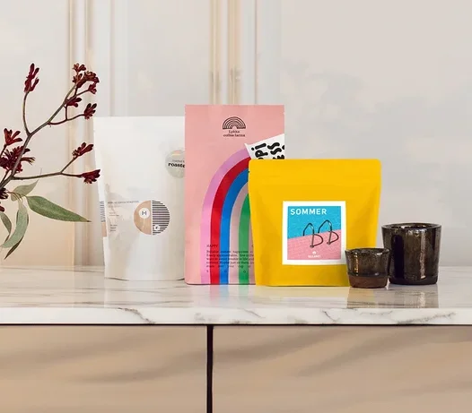 Drei bunte Produktbeutel in Weiß, Rosa mit Regenbogen und Gelb stehen auf einer Marmorarbeitsplatte neben zwei Vasen mit roten Blumen und zwei dunklen Tassen.