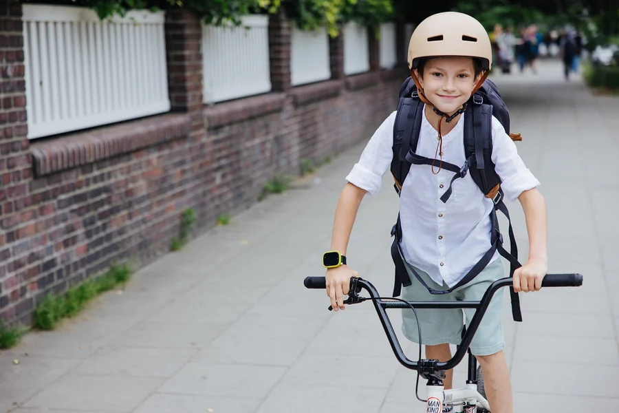 Junge mit Rucksack und beigem Helm fährt mit dem Fahrrad auf dem Gehweg.