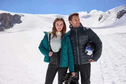 Mann und Frau stehen in einer verschneiten Berglandschaft. Die Frau trägt eine türkise Steppjacke und schwarze Skihose und hält Handschuhe. Der Mann trägt eine dunkle Steppjacke und hält einen Skihelm mit Skibrille.