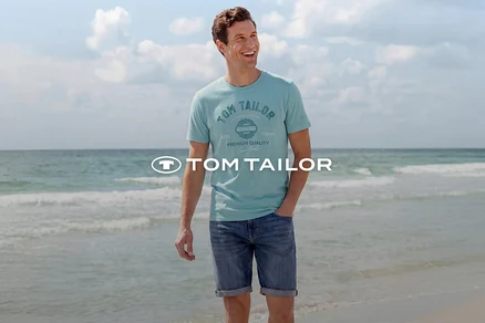 Lachender Mann in hellblauem T-Shirt mit Aufdruck und Jeans-Shorts steht am Strand vor dem Meer. Tom Tailor Logo.
