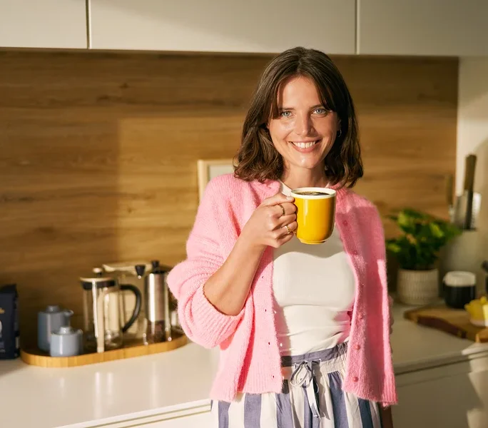 Lächelnde Frau hält eine gelbe Kaffeetasse in einer Küche. Sie trägt einen rosa Cardigan, ein weißes Oberteil und gestreifte Hosen. Auf der Theke hinter ihr stehen zwei French Press Kaffeebereiter, ein blaues Zucker- und Milchset sowie eine blaue Kaffeepackung.