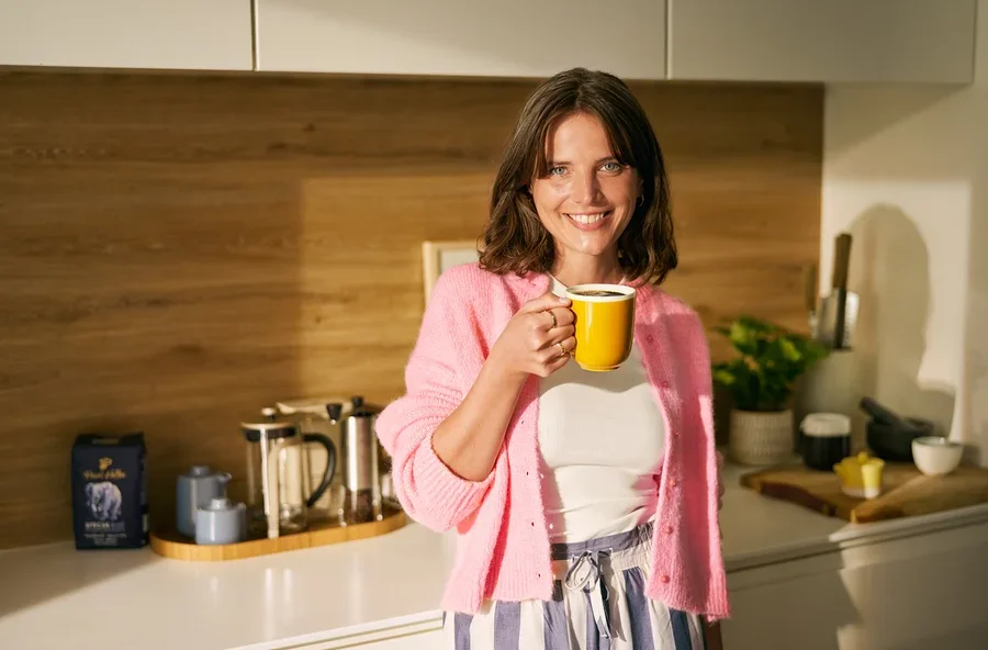 Lächelnde Frau hält eine gelbe Kaffeetasse in einer Küche. Sie trägt einen rosa Cardigan, ein weißes Oberteil und gestreifte Hosen. Auf der Theke hinter ihr stehen zwei French Press Kaffeebereiter, ein blaues Zucker- und Milchset sowie eine blaue Kaffeepackung.