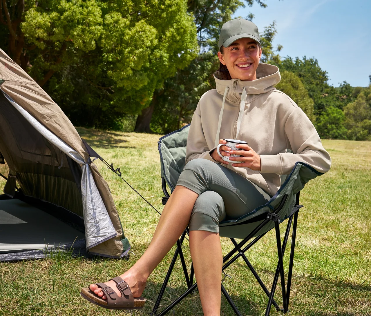 Frau sitzt im Tambu »ASANA« Camping-Faltstuhl, hält eine Tasse und trägt 3/4-Outdoor-Leggings und einen Kapuzenpullover.