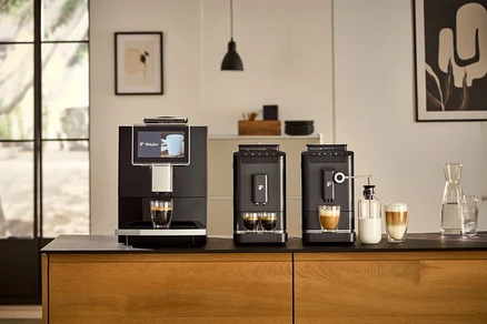 Drei »Esperto2 Caffè« Kaffeevollautomaten in verschiedenen Farben stehen nebeneinander auf einer Küchentheke. Zwei Tassen sind mit Kaffee und eine Tasse mit Milch gefüllt. Im Hintergrund befindet sich eine Milchkaraffe.