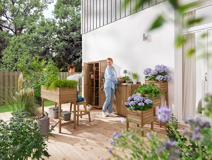 Ein Junge und eine Frau hantieren auf einer hölzernen Terrasse mit Gartenmöbeln und Pflanzen.