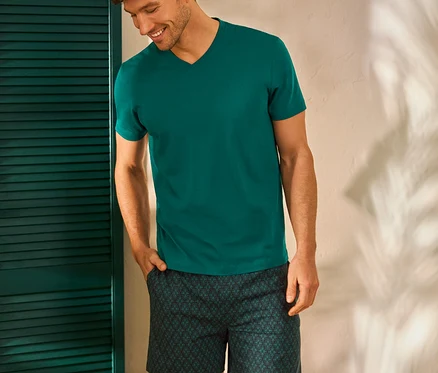 Lächelnder Mann mit Hand in der Hosentasche trägt ein grünes T-Shirt und Shorts mit Muster, stehend neben einem Hocker mit Büchern.