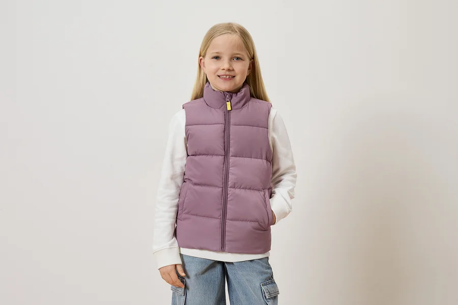 Mädchen mit blonden Haaren trägt eine lila Weste über einem weißen Pullover und blauen Jeans mit Taschen.