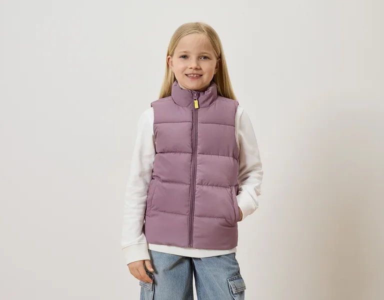 Mädchen mit blonden Haaren trägt eine lila Weste über einem weißen Pullover und blauen Jeans mit Taschen.