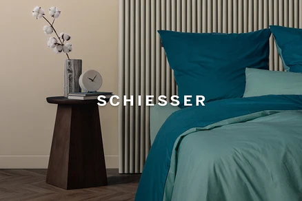 Ein Schlafzimmer mit einem Holztisch, auf dem eine Vase mit Baumwollzweigen, eine Uhr und Bücher stehen. Rechts ein Bett mit türkisfarbener und hellgrüner Bettwäsche.