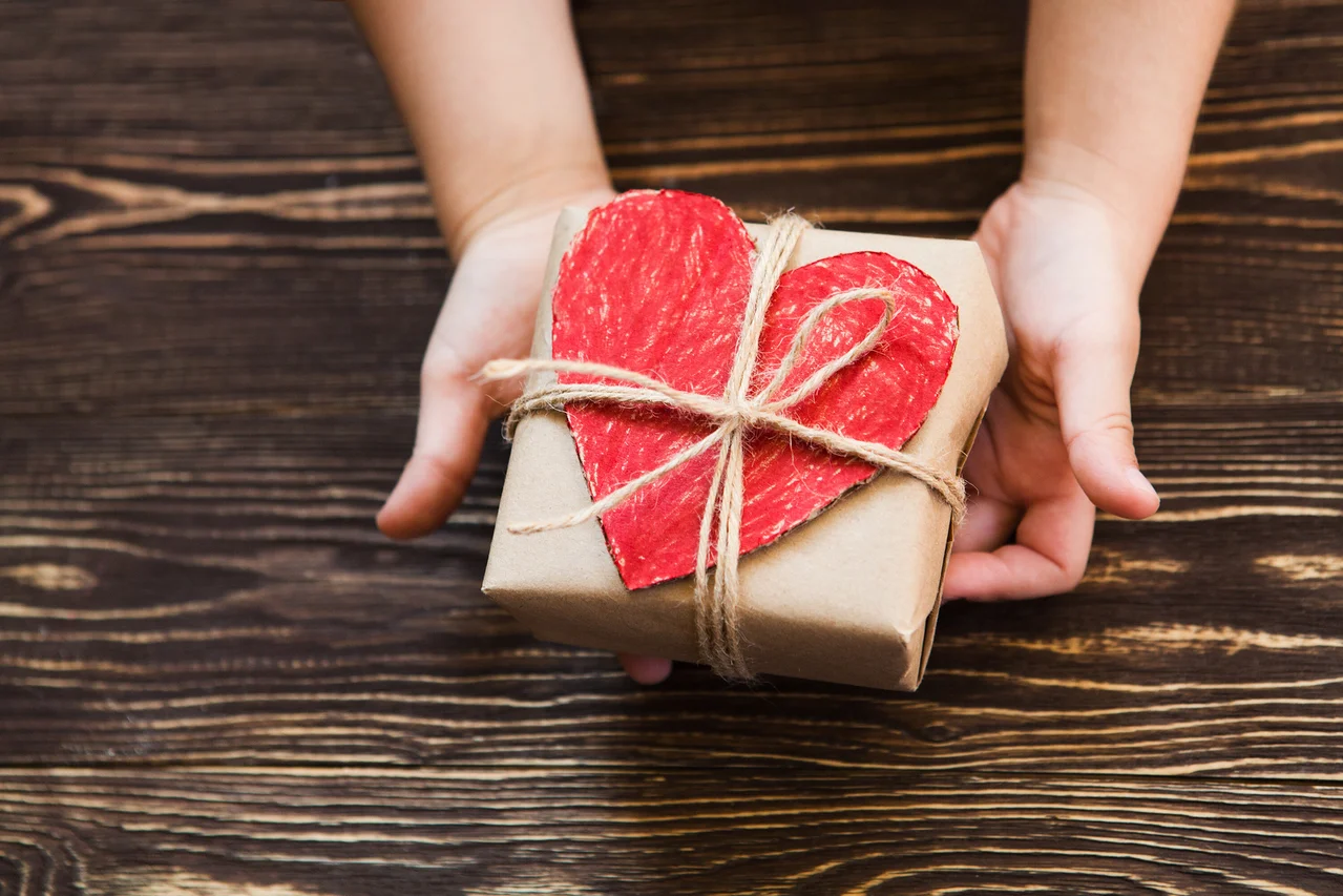 Hände halten ein braunes Geschenk mit einem roten Herz, das mit einer Schnur auf einem Holzuntergrund befestigt ist.