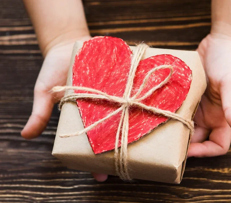 Hände halten ein braunes Geschenk mit einem roten Herz, das mit einer Schnur auf einem Holzuntergrund befestigt ist.