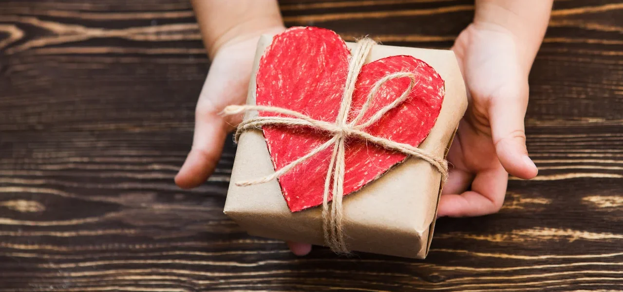 Hände halten ein braunes Geschenk mit einem roten Herz, das mit einer Schnur auf einem Holzuntergrund befestigt ist.