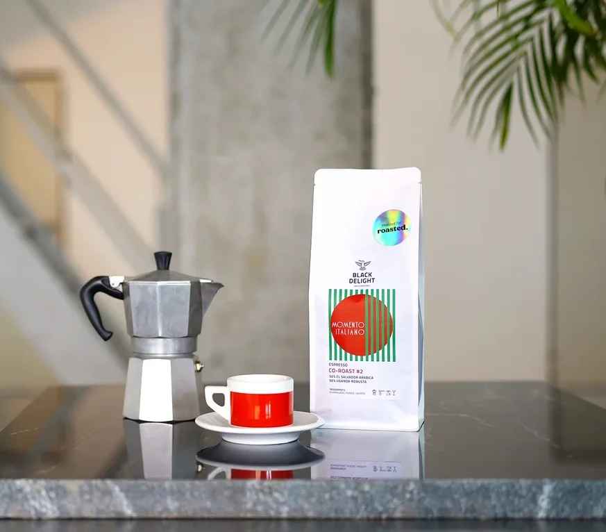 Auf einem Tisch stehen eine Kaffeemaschine, eine Tasse und eine Packung Black Delight x roasted. - Momento Italiano Co-Roast #2 Espresso.