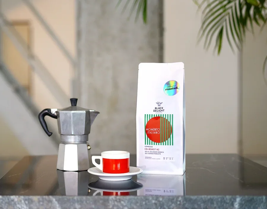Auf einem Tisch stehen eine Kaffeemaschine, eine Tasse und eine Packung Black Delight x roasted. - Momento Italiano Co-Roast #2 Espresso.