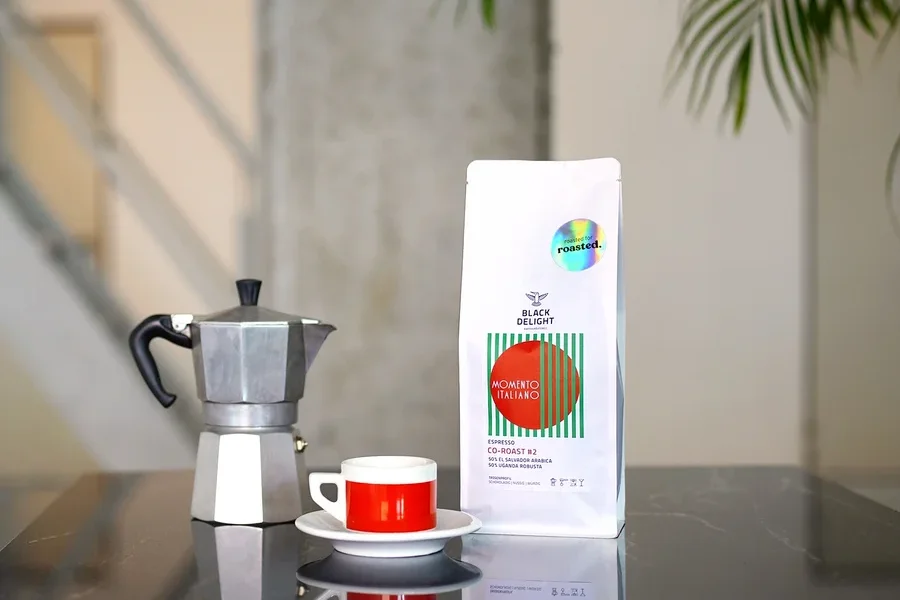 Auf einem Tisch stehen eine Kaffeemaschine, eine Tasse und eine Packung Black Delight x roasted. - Momento Italiano Co-Roast #2 Espresso.