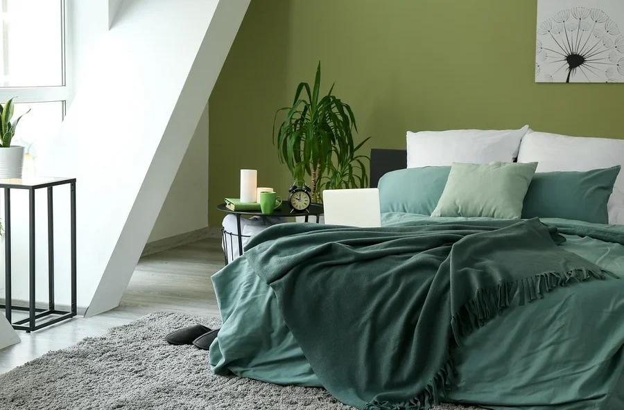 Schlafzimmer mit grün gestrichener Wand, Bett mit grüner Decke und Kissen, Beistelltisch mit Laptop, Tasse und Uhr.