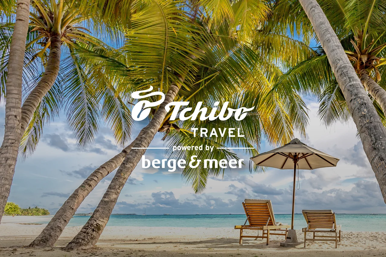 Zwei Liegestühle und ein Sonnenschirm stehen am Strand unter Palmen. Im Vordergrund befindet sich das Tchibo TRAVEL Logo.