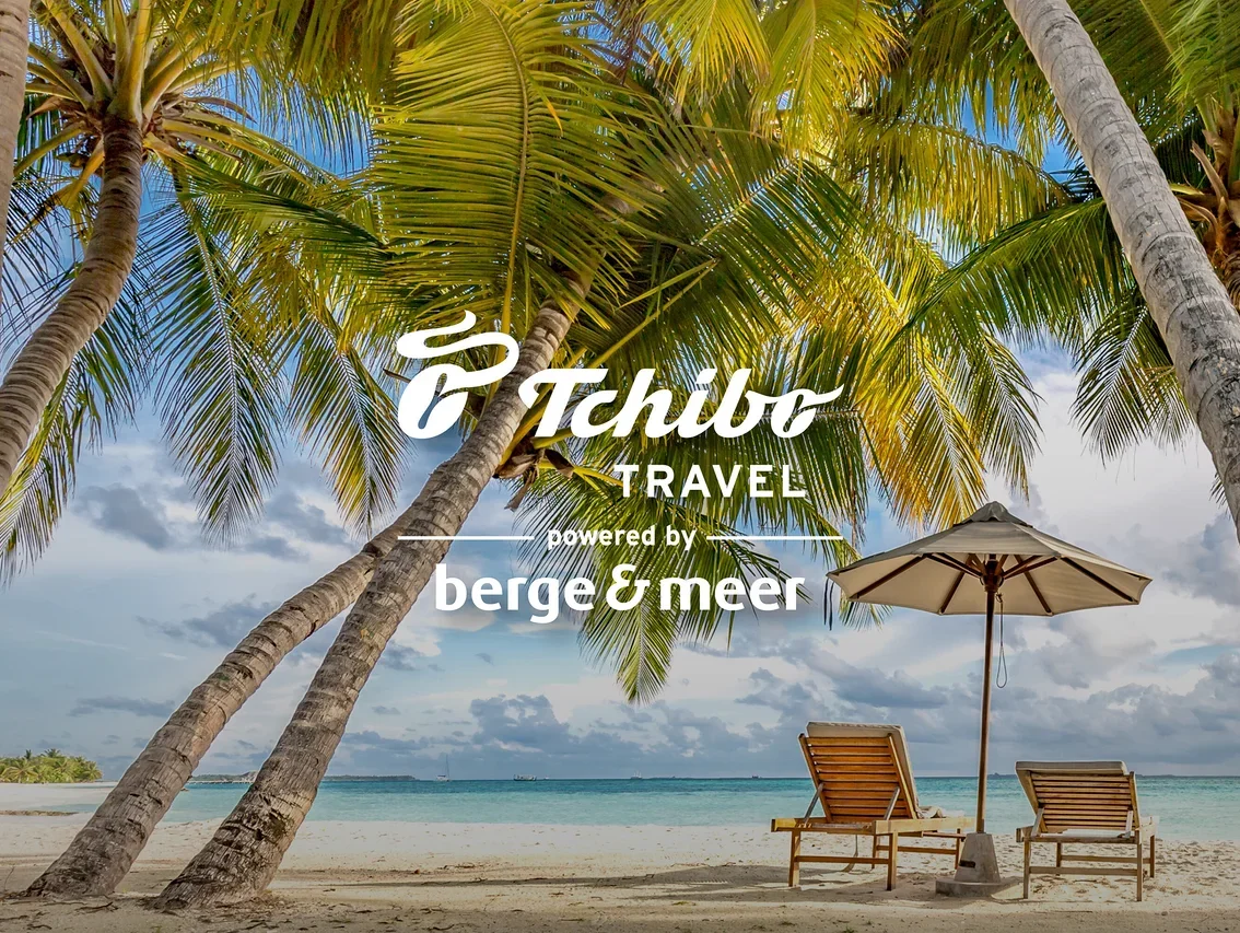 Zwei Liegestühle und ein Sonnenschirm stehen am Strand unter Palmen. Im Vordergrund befindet sich das Tchibo TRAVEL Logo.