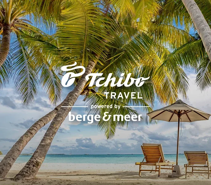 Zwei Liegestühle und ein Sonnenschirm stehen am Strand unter Palmen. Im Vordergrund befindet sich das Tchibo TRAVEL Logo.