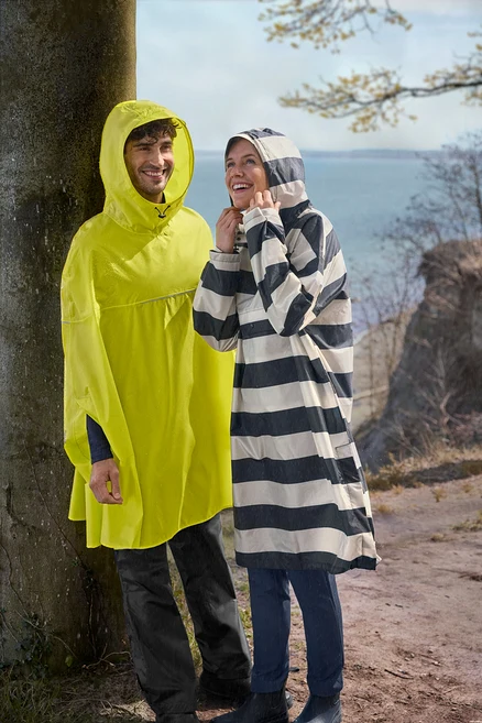 Mann in gelbem Regenponcho und Frau in schwarz-weiß gestreiftem Regenponcho lächeln im leichten Regen im Freien nahe einem Baum und Wasser.