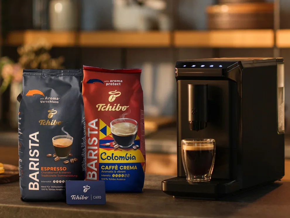 Zwei Packungen Tchibo Barista Kaffee, Espresso und Colombia Caffè Crema, stehen neben einer schwarzen Kaffeemaschine mit einer Tasse Kaffee und einer Tchibo Card auf einer Küchenzeile.
