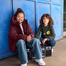 Zwei Kinder sitzen vor einer blauen Wand. Ein Mädchen in einer bordeauxroten Pufferjacke lächelt links, während ein Junge in einem grauen Hoodie mit Smiley auf einer Boombox rechts sitzt.