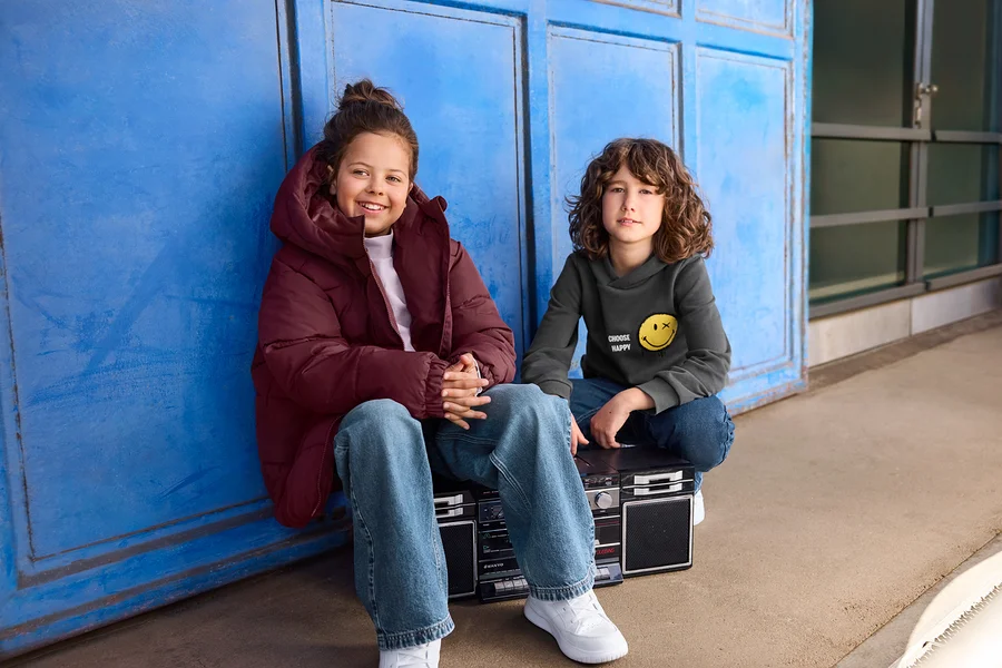 Zwei Kinder sitzen vor einer blauen Wand. Ein Mädchen in einer bordeauxroten Pufferjacke lächelt links, während ein Junge in einem grauen Hoodie mit Smiley auf einer Boombox rechts sitzt.