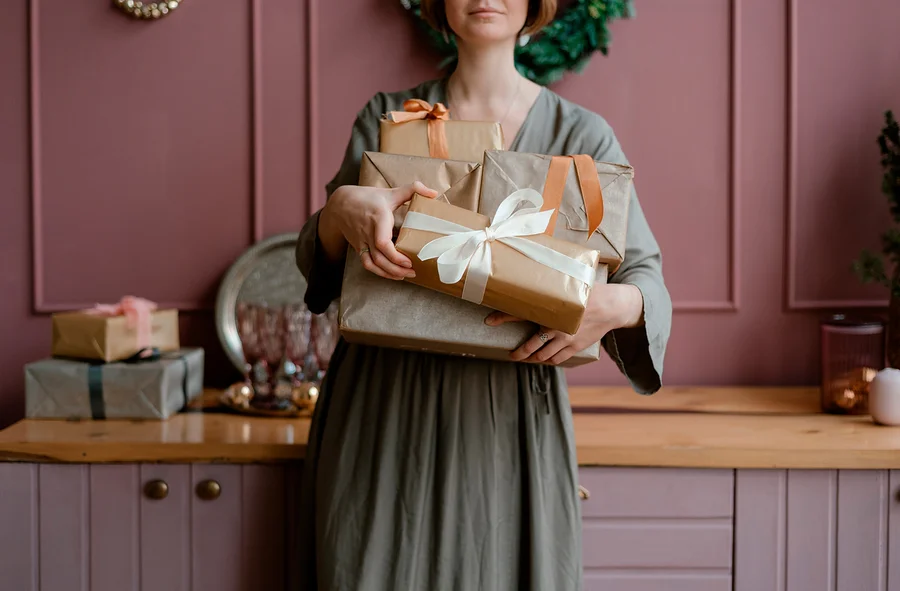 Eine Person in einem grauen Kleid hält einen Stapel Geschenke, die in braunes Papier mit weißen und orangefarbenen Bändern eingepackt sind. Im Hintergrund sind eine rosa Wand und eine Holztheke zu sehen.
