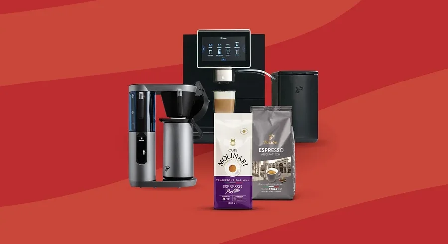 Eine schwarze Kaffeemaschine brüht Kaffee in ein Glas, daneben steht ein schwarzer Milchaufschäumer. Links befindet sich eine graue Filterkaffeemaschine. Davor liegen eine weiß-violette Molinari Kaffeepackung und eine graue Tchibo Espresso-Packung auf rotem Hintergrund.