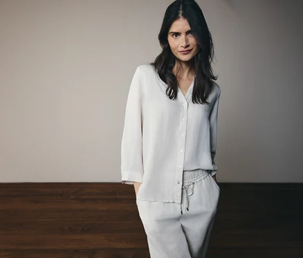 Frau mit langen, dunklen Haaren, die ein helles, zweiteiliges Pyjama-Set aus Hemd und Hose trägt und in die Hosentaschen greift.