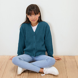 Ein Mädchen sitzt mit gekreuzten Beinen auf einem Holzboden, gekleidet in eine blaue Strickjacke, eine weiße Bluse, Jeans und weiße Turnschuhe.