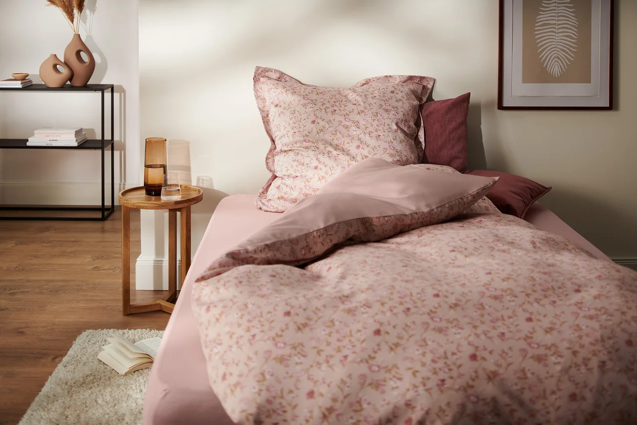 Schlafzimmer mit Bettwäsche-Set aus rosafarbenem Satin mit Blumenmuster, Nachttisch und Dekorationen. Im Bild sind eine Satin-Bettwäsche, Übergröße und eine Satin-Bettwäsche, Normalgröße zu sehen.