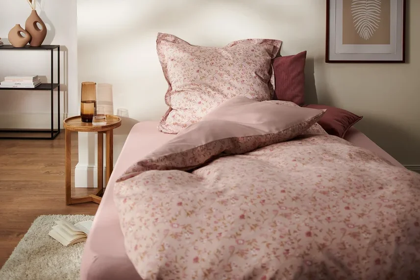 Schlafzimmer mit Bettwäsche-Set aus rosafarbenem Satin mit Blumenmuster, Nachttisch und Dekorationen. Im Bild sind eine Satin-Bettwäsche, Übergröße und eine Satin-Bettwäsche, Normalgröße zu sehen.